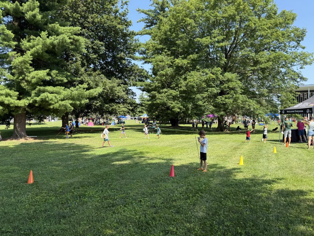 Children on a green lawn with large trees looming above. Cones and rope are placed strategically as the children practice following the rope with their hands with eyes closed.