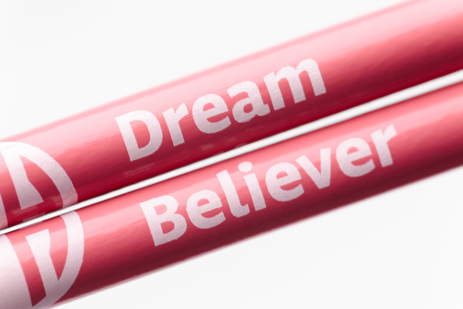 Two pomegranate pencils held closely together to show the full black APH logo. The pencil on top says "Dream" and the pencil on bottom says "Believer."