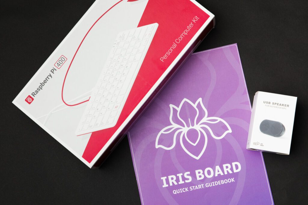 The purple Iris Board user guide on a black background next to boxes containing a speaker and a pink Raspberry Pi keyboard.